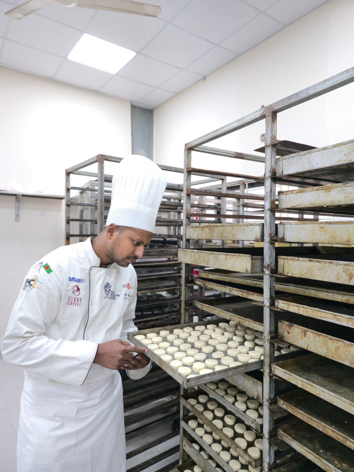 Chef Munna inspecting trays in a production kitchen surrounded by rack ovens and sheet pans